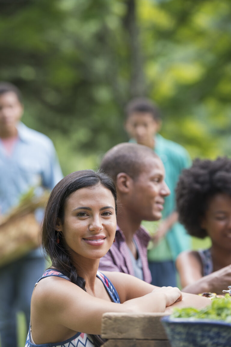A woman smiles at the camera. She is sat at a picnic table outside, there are other people also at the table.