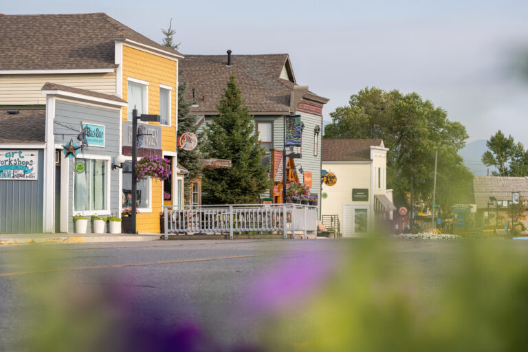 Colourful storefronts line the street in downtown. It is a warm, hazy day and a mountain pokes through in the background. Purple flowers sit out of focus in the foreground. There are large conifers between the buildings.