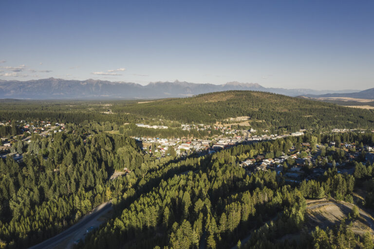 An areal shot of the community on a bright sunny day. The river flows through the center of the image, with homes on either side, tucked away amongst trees. Dense conifer forest surrounds the town, which sits in a bowl of mountains. Fischer Peak rises in the background.