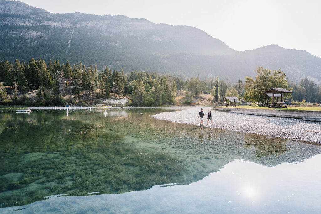 Two people stroll along the beach at Tilley Memorial Park on the shore of Columbia Lake. The emerald green water is very still and clear, so rocks are visible at the bottom of the lake. A dock is visible on the lake. Conifers line the lake, and large mountains rise behind them.