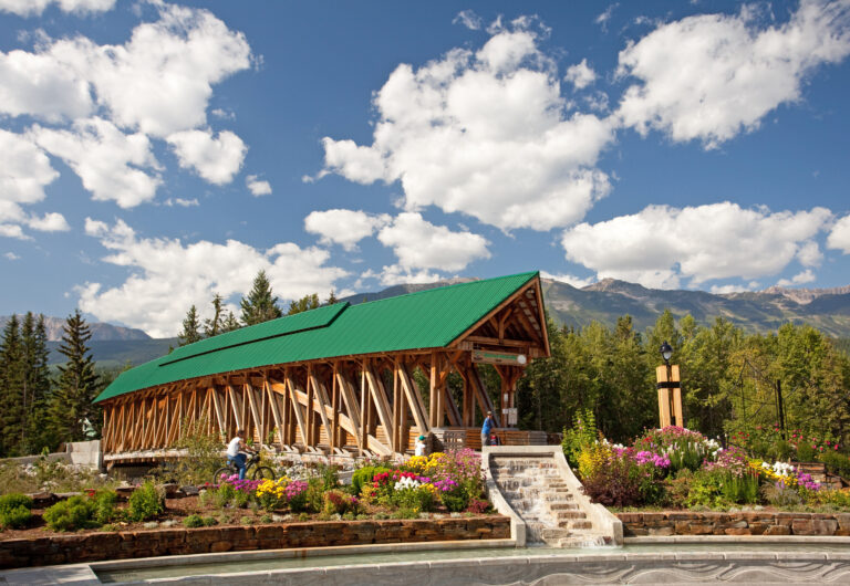 A photo of the Kicking Horse Pedestrian Bridge spanning the river on a bright, sunny day. There are blooming flower beds by the entrance to the bridge and bright mountains in the background.